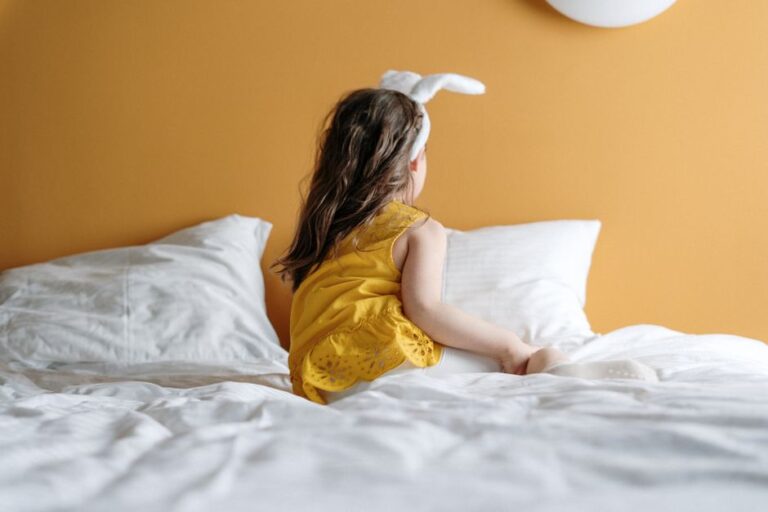 A young girl in bunny ears enjoys a cozy moment on a bed with a warm yellow background.