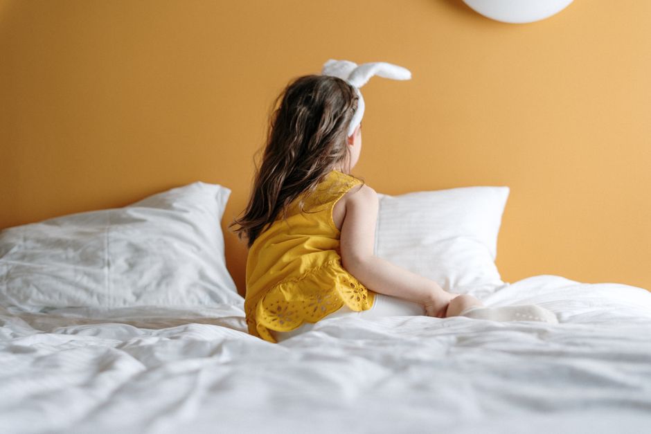 A young girl in bunny ears enjoys a cozy moment on a bed with a warm yellow background.